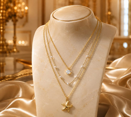 A gold-tone layered necklace set displayed on a cream-colored jewelry bust. The set includes a delicate chain with a small clear crystal pendant, a textured chain with tiny pearl-like beads, and a longer chain featuring a gold starfish pendant.