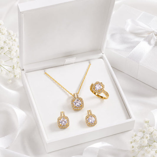 A gold jewelry set with a pendant necklace, ring, and two matching earrings displayed inside an open white gift box. The pieces feature clear gemstones surrounded by gold detailing, with white satin ribbons and small white flowers arranged around the box.
