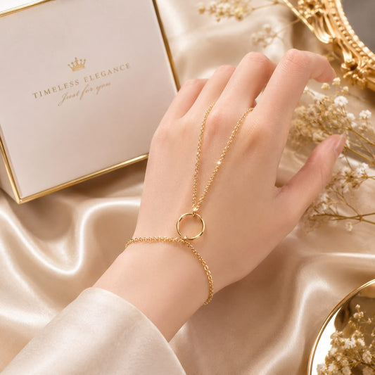 A gold chain bracelet with a double ring design worn on a woman's wrist, styled on a beige satin background with a branded 'TIMELESS ELEGANCE' jewelry box and decorative dried flowers.
